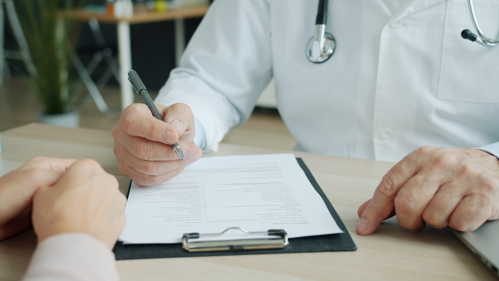 Le mani di un medico che compila a penna un modulo su una cartellina, con le mani della paziente appoggiate al tavolo dall'altra parte.